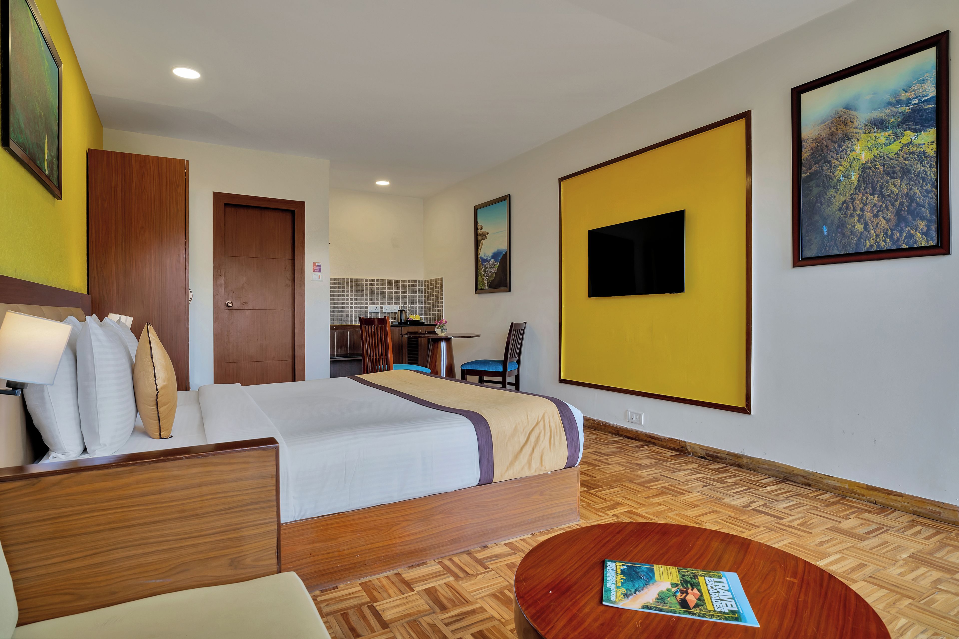 Quarto de hotel com cama, escrivaninha, cadeira, mesa de madeira redonda e televisão na parede.