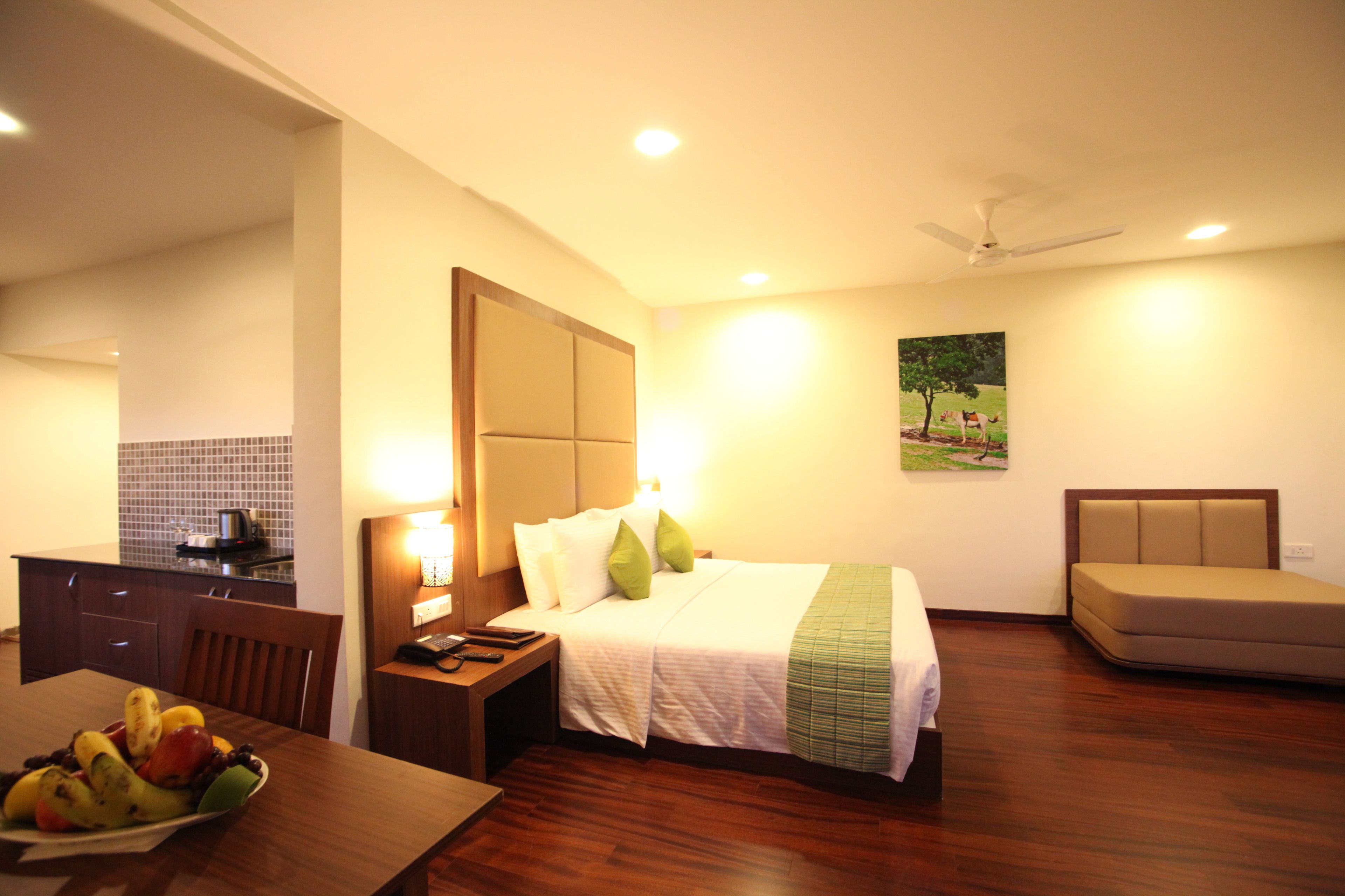 A hotel room with a bed, a bedside table with a lamp, a chair, a small table with fruit, and a painting on the wall.