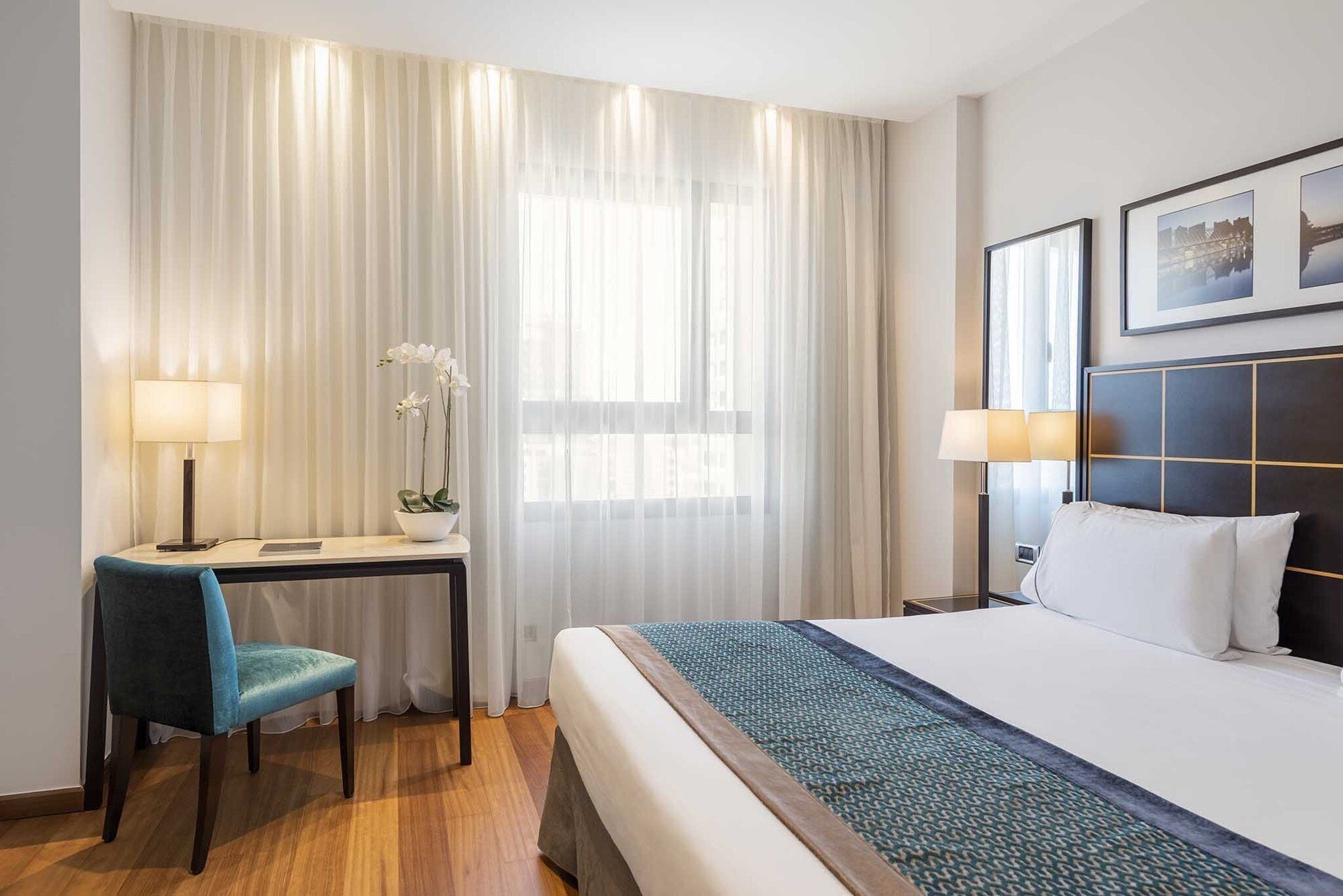 A hotel room with a bed, a desk, and a chair. There are two lamps on either side of the bed.
