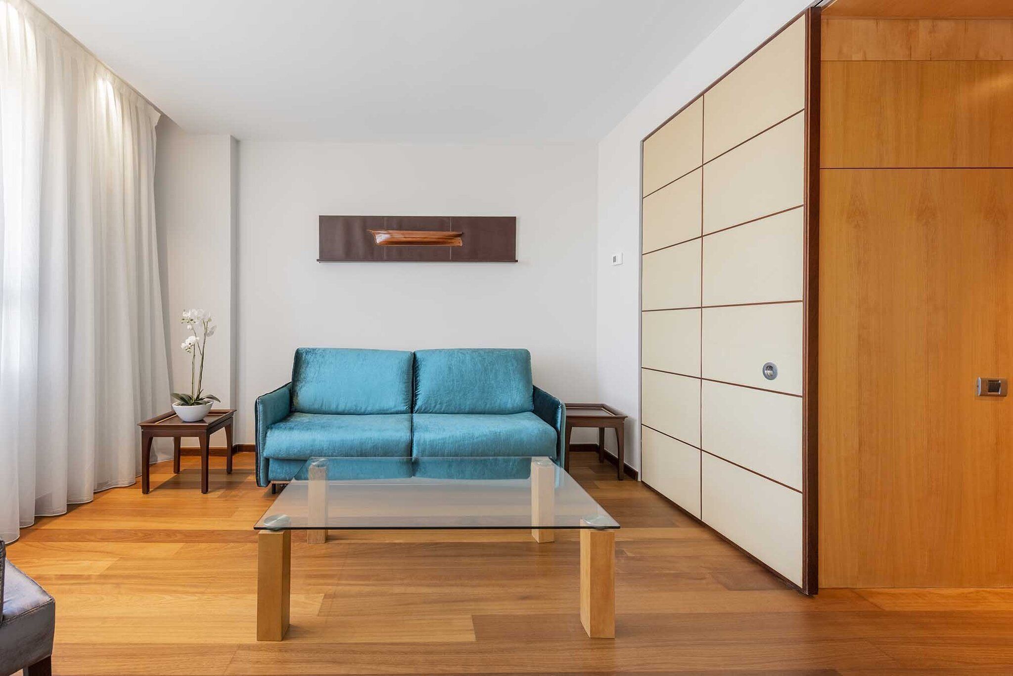 Una sala moderna con un sofá azul, una mesa de centro de cristal, suelo de madera, un armario de madera y una pared blanca con un cuadro enmarcado.