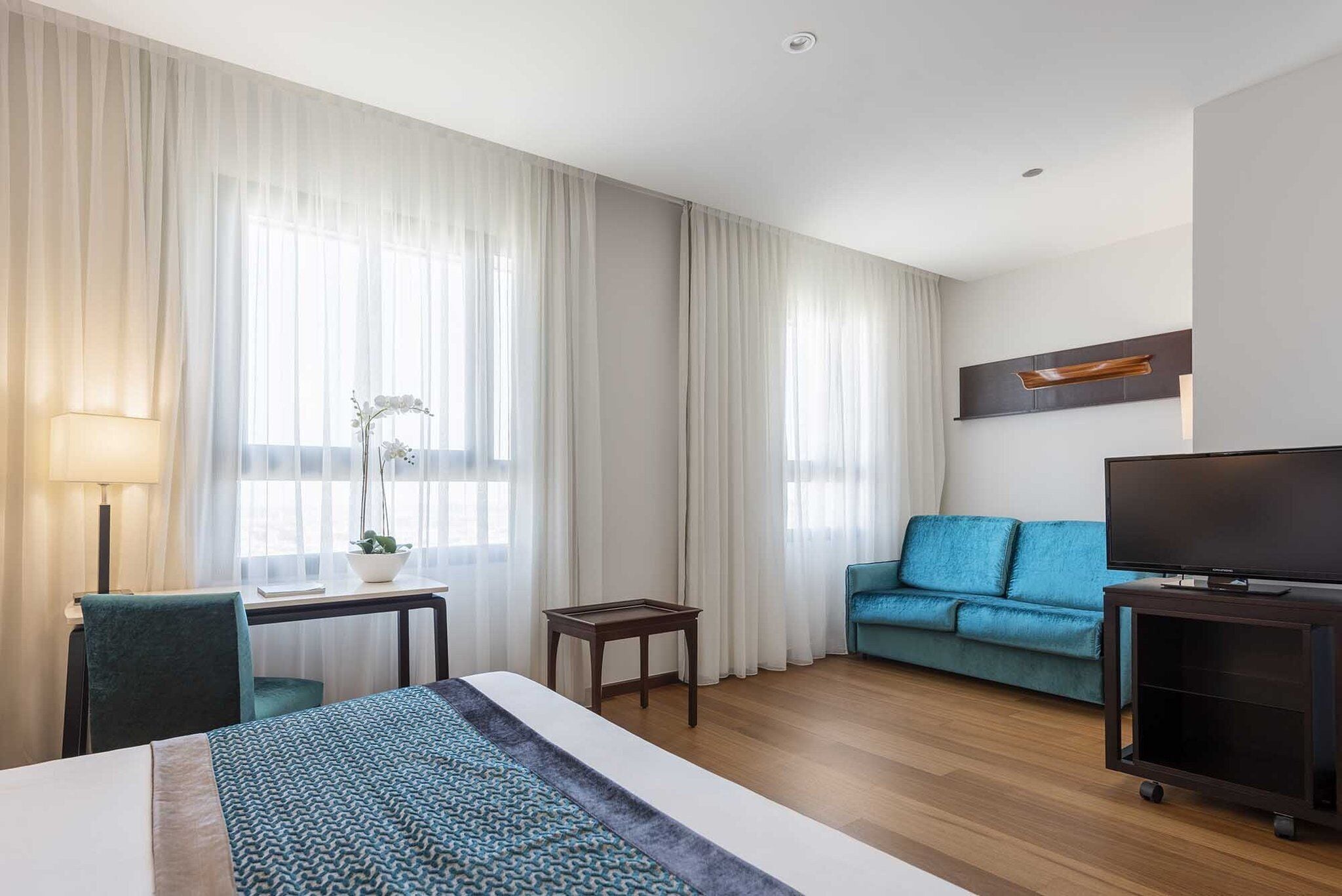 Habitación de hotel con cama, escritorio, silla, sofá, televisor y ventana con cortinas.