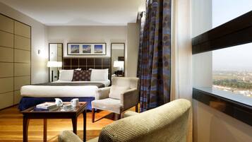 Habitación de hotel con una cama grande, dos sillones, una mesita con un libro y una taza, y vistas a la ciudad desde la ventana.