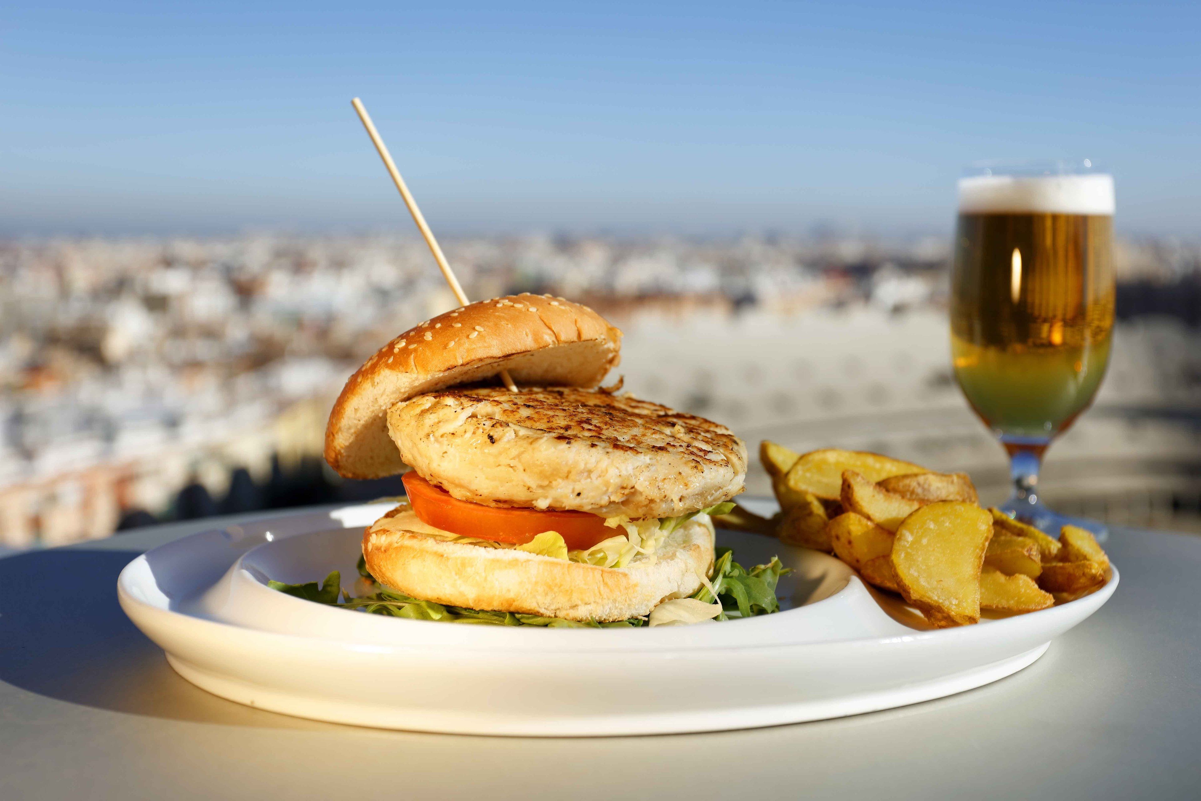 Un plato con un sándwich a la parrilla, papas fritas y una cerveza en una mesa, con vistas a la ciudad de fondo.