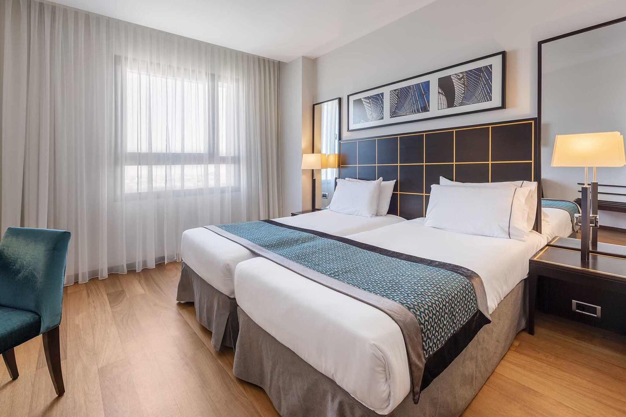 A hotel room with a large bed, two bedside tables, a chair, and a window with sheer curtains.
