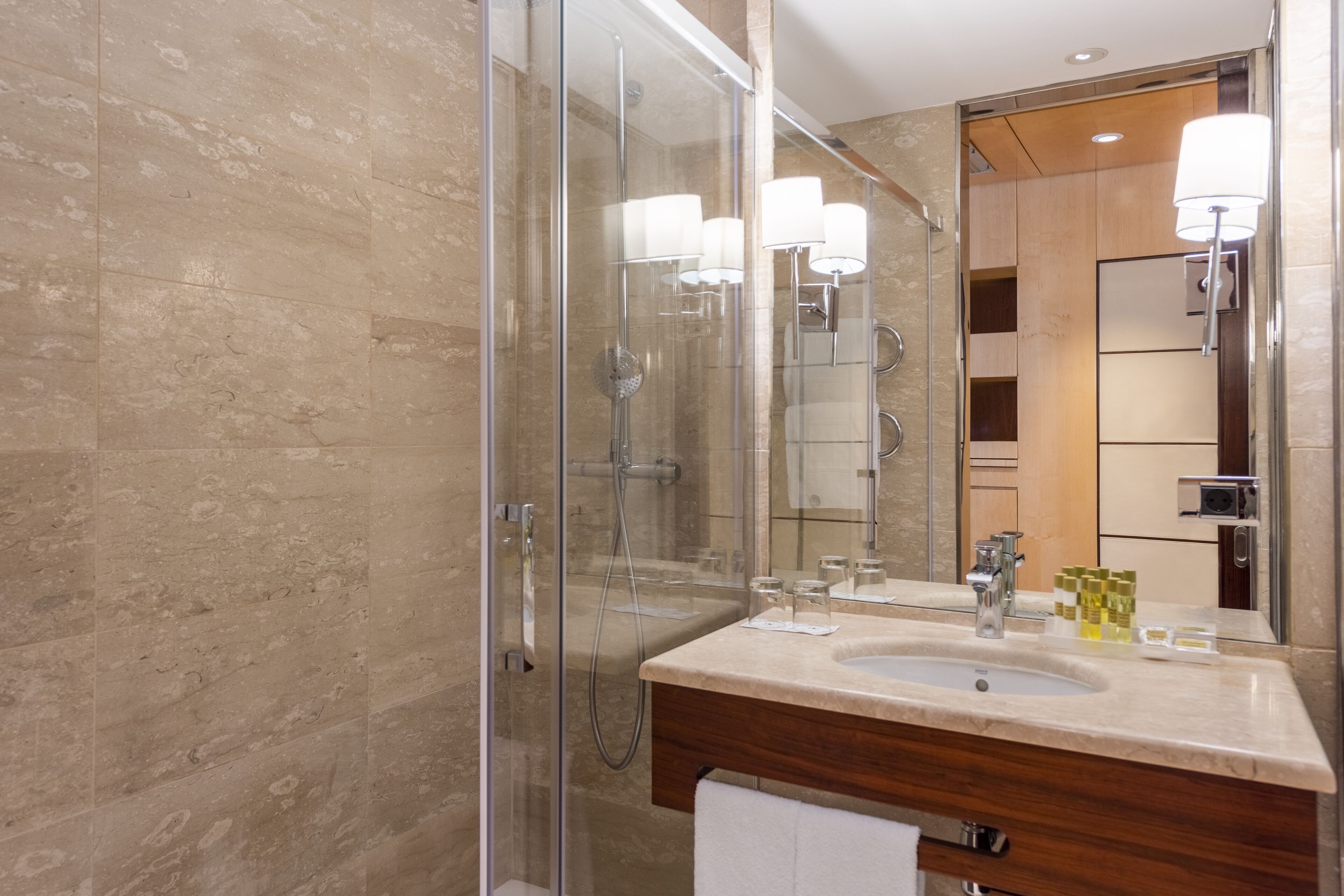 Un baño moderno con ducha envuelta en vidrio, tocador de madera con lavabo y un espejo grande con luces empotradas.