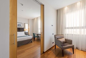 Ein Hotelzimmer mit einem Bett, einem Schreibtisch, einem Sessel und einem großen Fenster mit durchsichtigen Vorhängen.