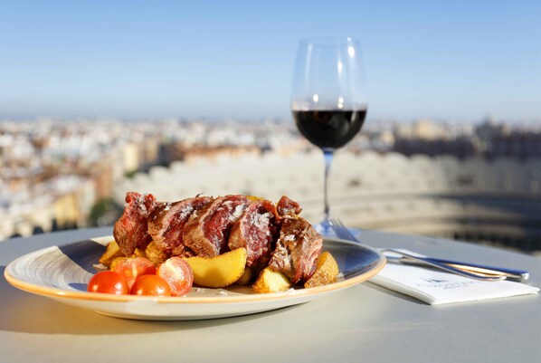 Une assiette remplie de viande et de pommes de terre, un verre de vin rouge et, en arrière-plan, une vue sur le paysage urbain.