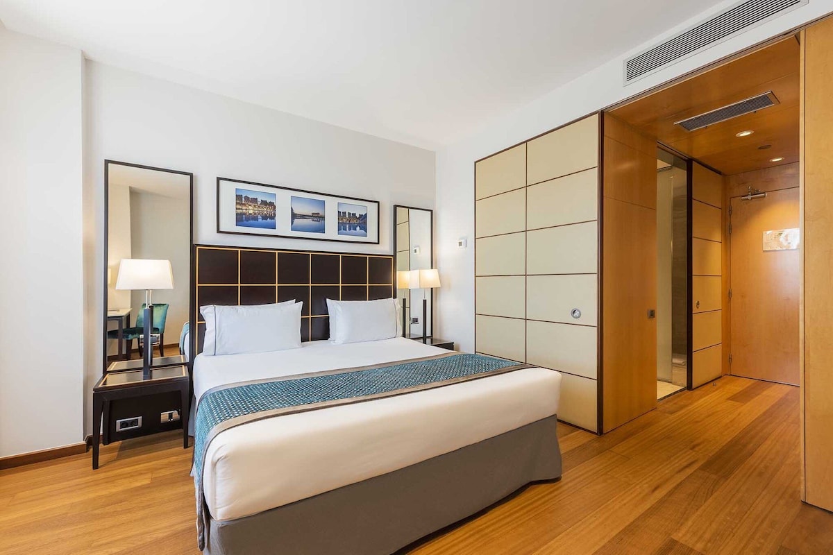 A hotel room with a large bed, two bedside tables with lamps, a large mirror, and a wardrobe with sliding doors.