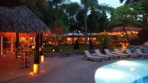 Un área de piscina en un resort, con sillas de descanso, una estructura de techo de paja y mobiliario exterior.