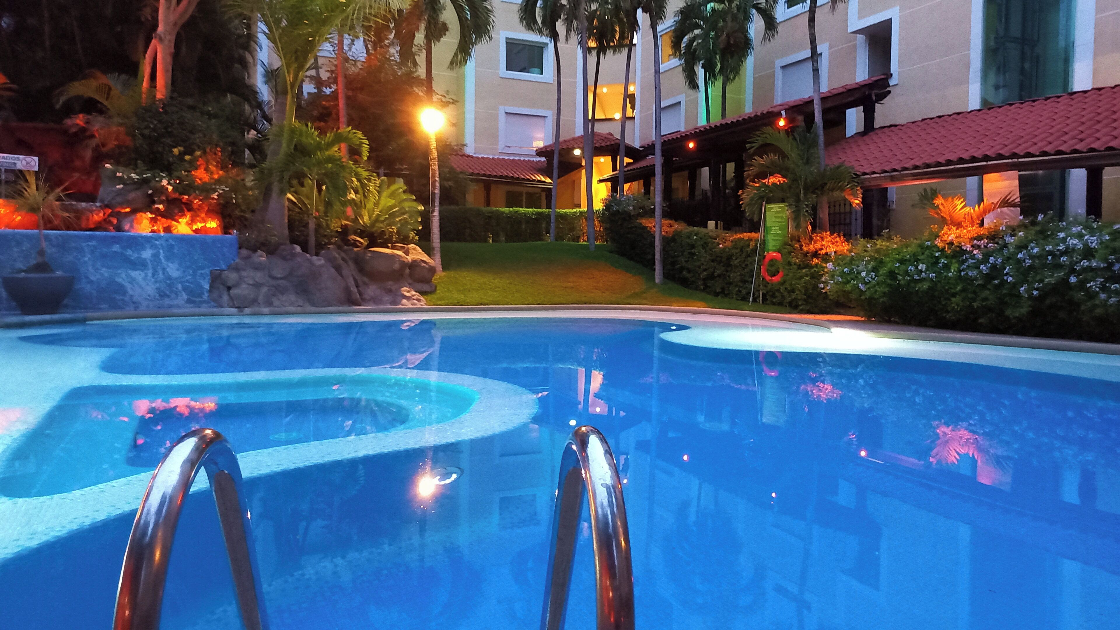 Una piscina con superficie de agua cristalina, rodeada por un jardín ajardinado con palmeras y un edificio con tejado de tejas rojas al fondo.