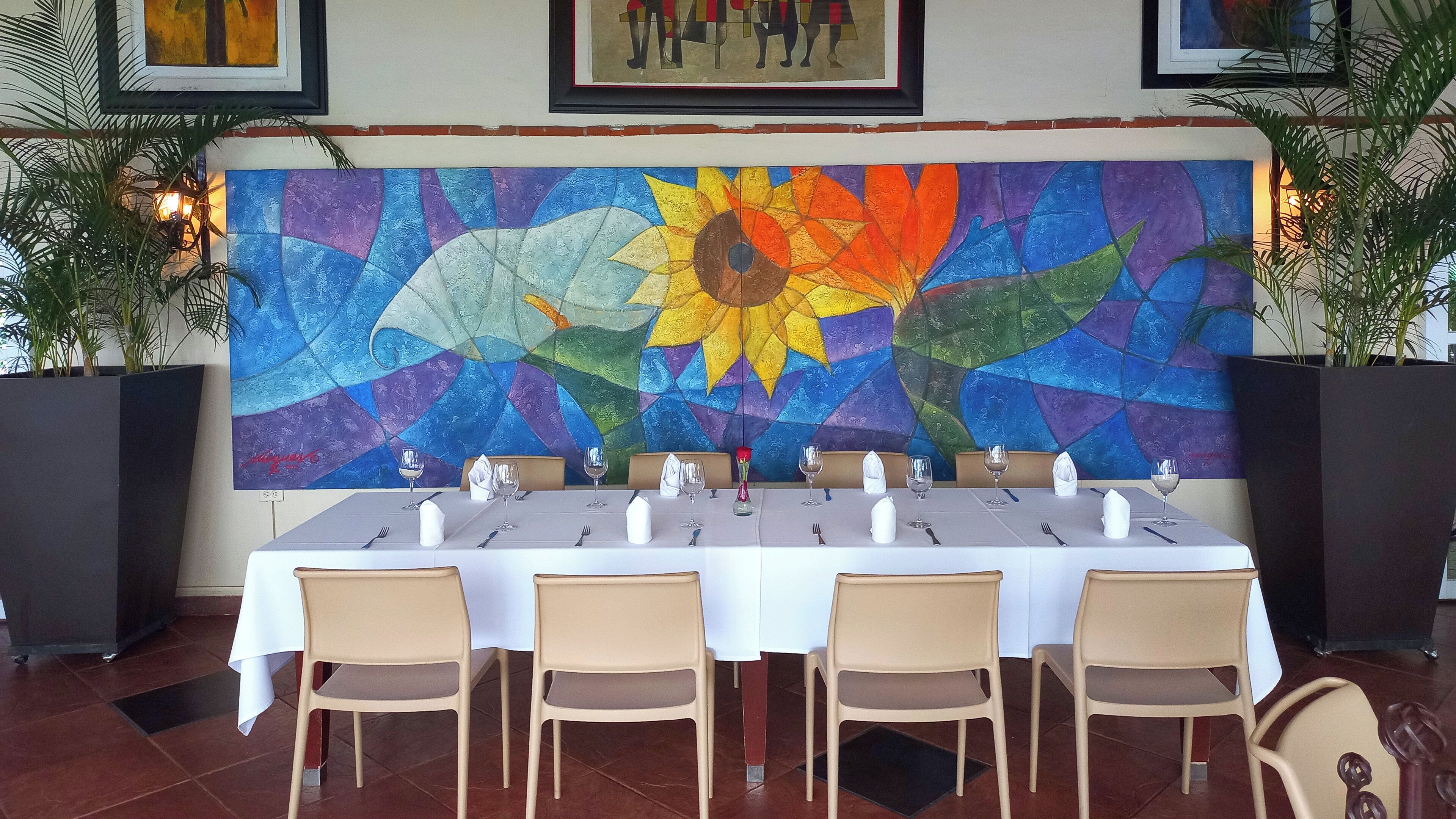 A dining area with a long table set for six, surrounded by chairs, with a colorful abstract mural on the wall behind.