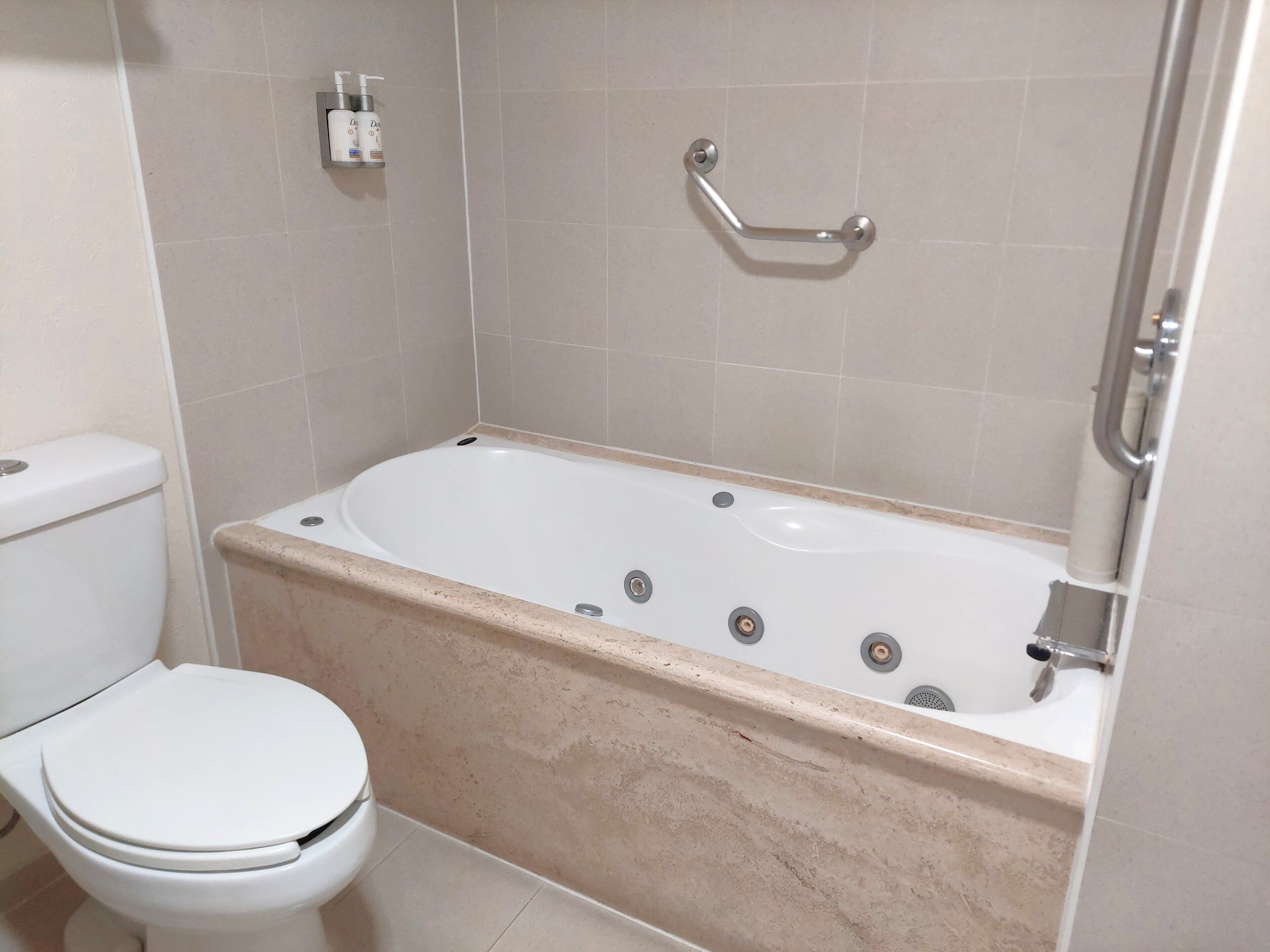 Un baño con un inodoro blanco, una bañera con un revestimiento de mármol color beige y una barra de apoyo sobre la bañera.