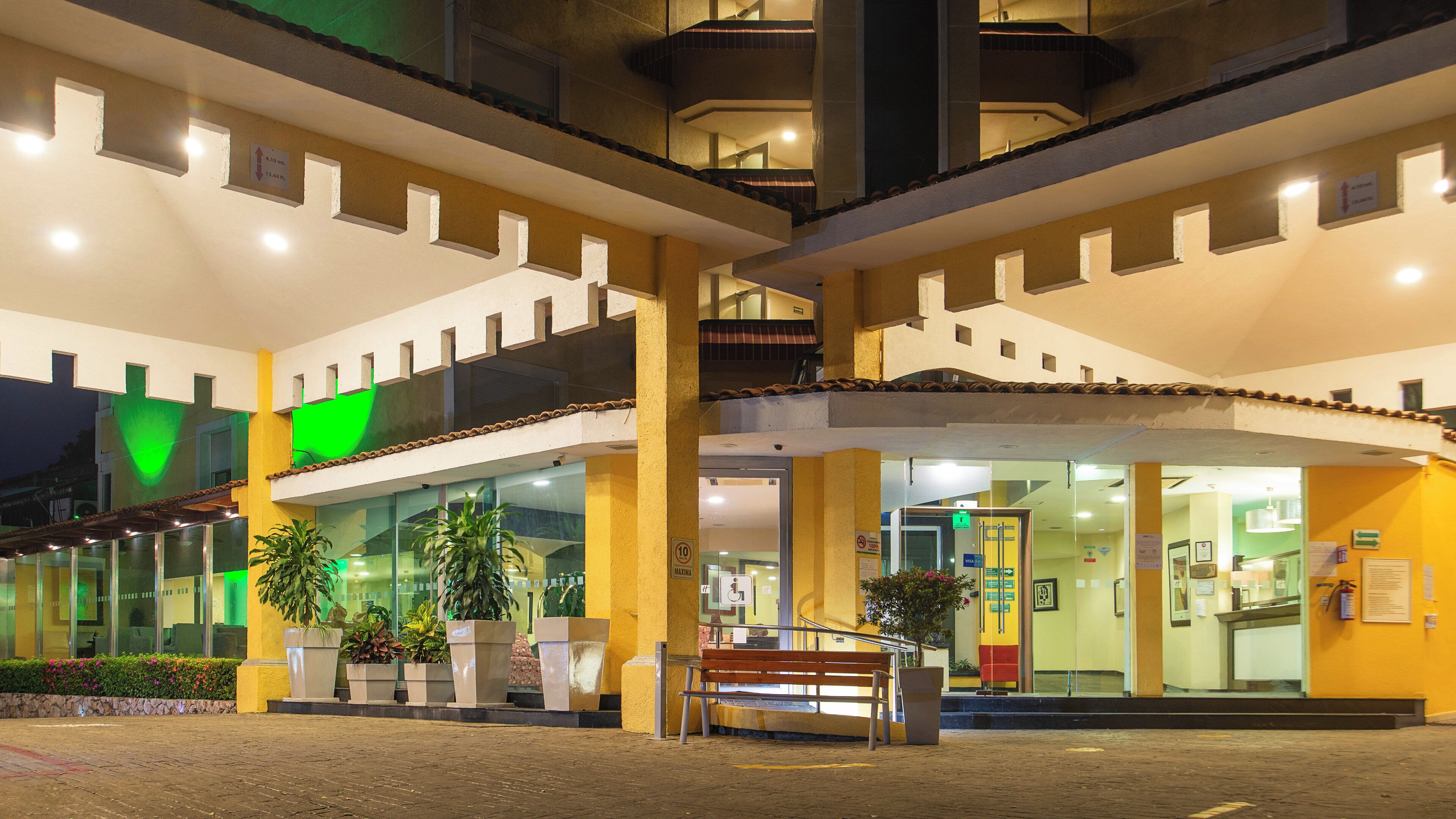 Entrada de hotel con rampa, plantas en macetas y un banco.