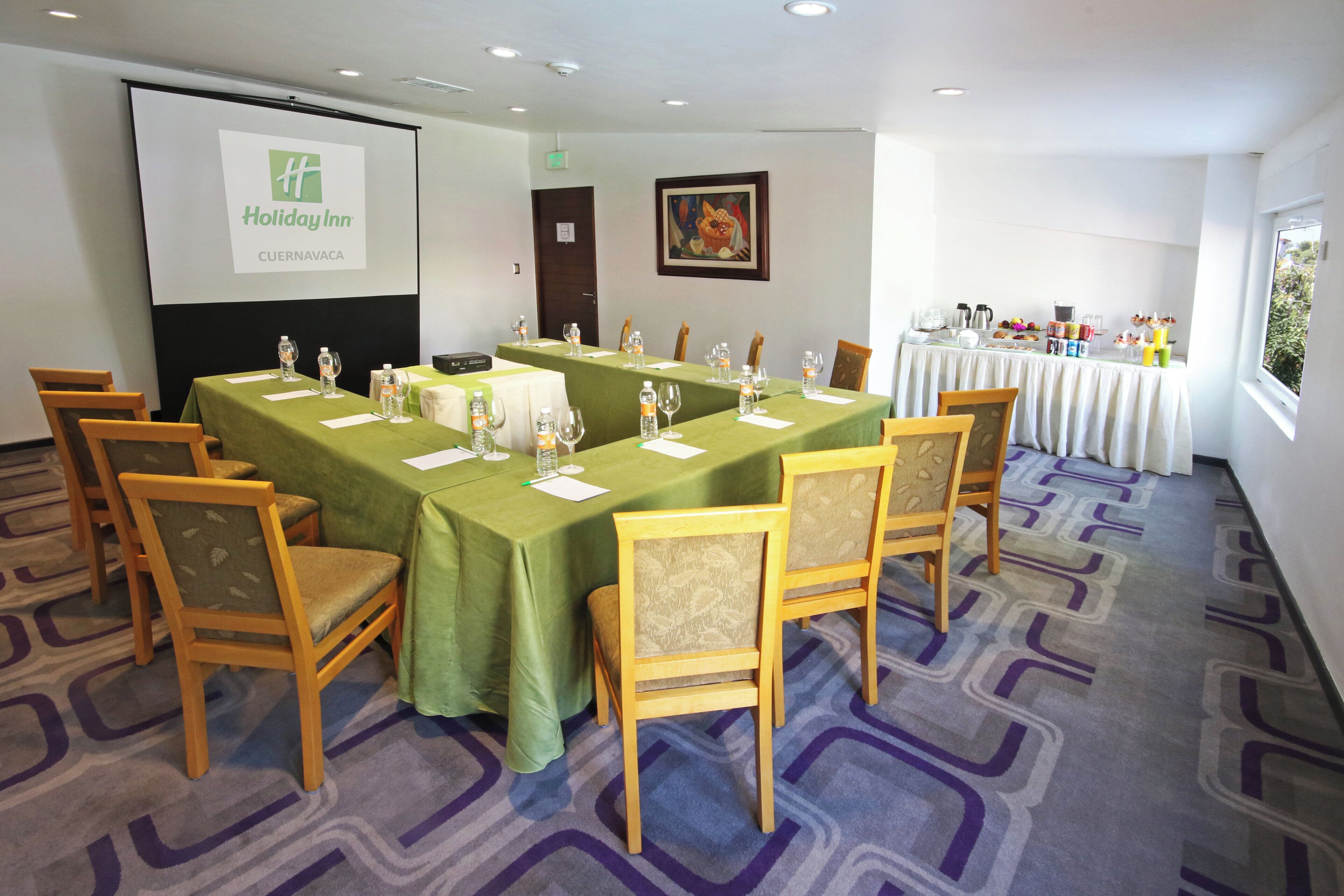 Ein Konferenzraum mit einer großen Leinwand, die das Holiday Inn Logo zeigt, grünen Tischdecken, Holzsesseln und einem Buffet mit Speisen und Getränken.
