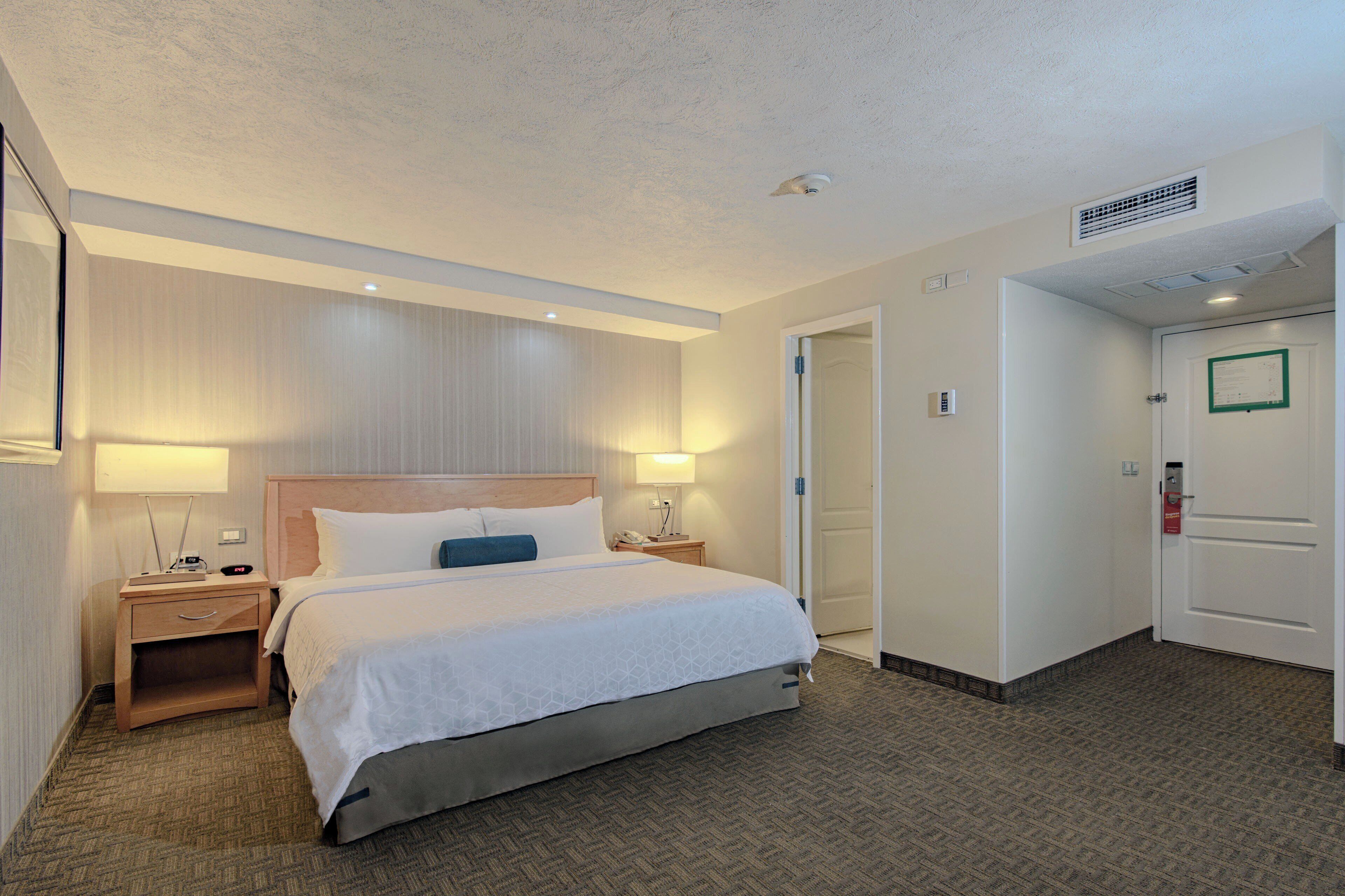A hotel room with a large bed, bedside tables, a nightstand, a wall-mounted lamp, and a door with a green sign.