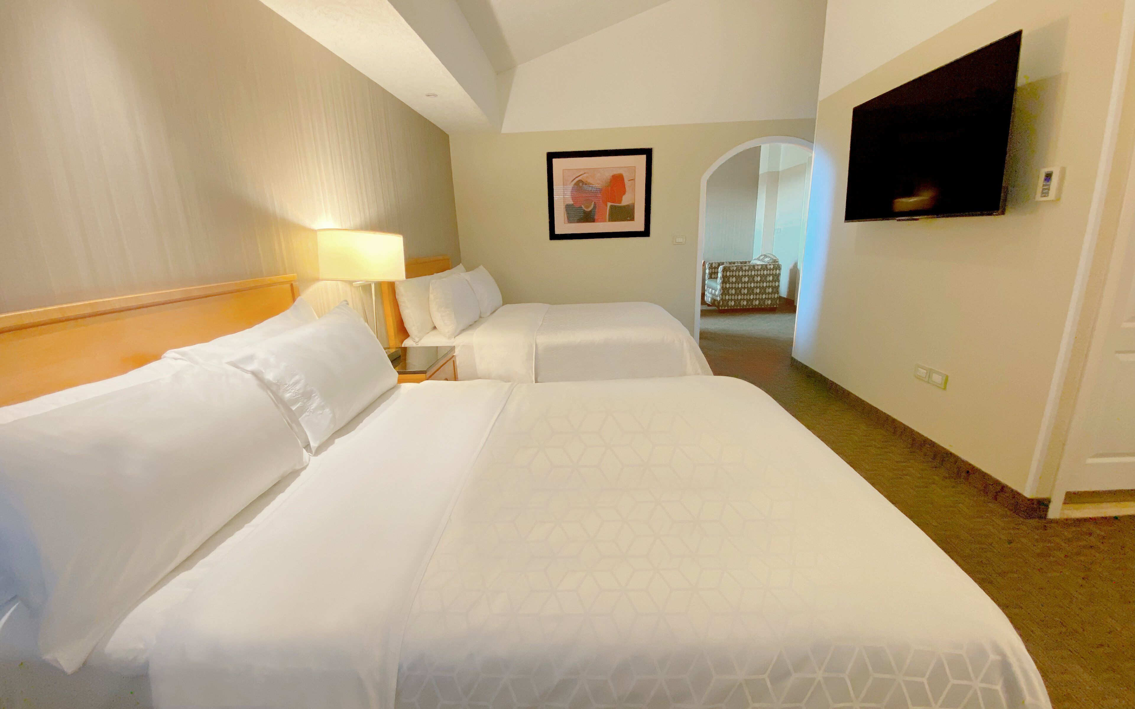 Quarto de hotel com duas camas, televisão, quadro emoldurado e corredor que dá para outro quarto.