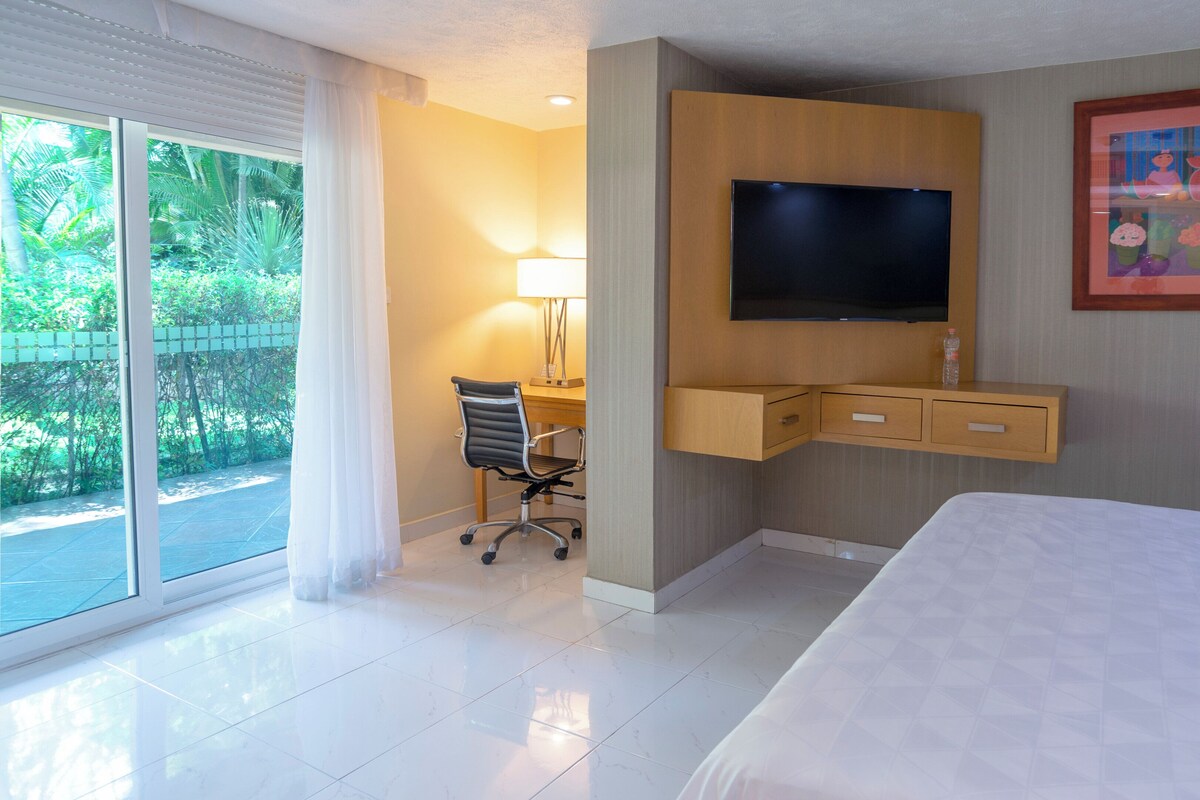 A hotel room with a bed, a desk with a chair, a television, and a window with a view of greenery.