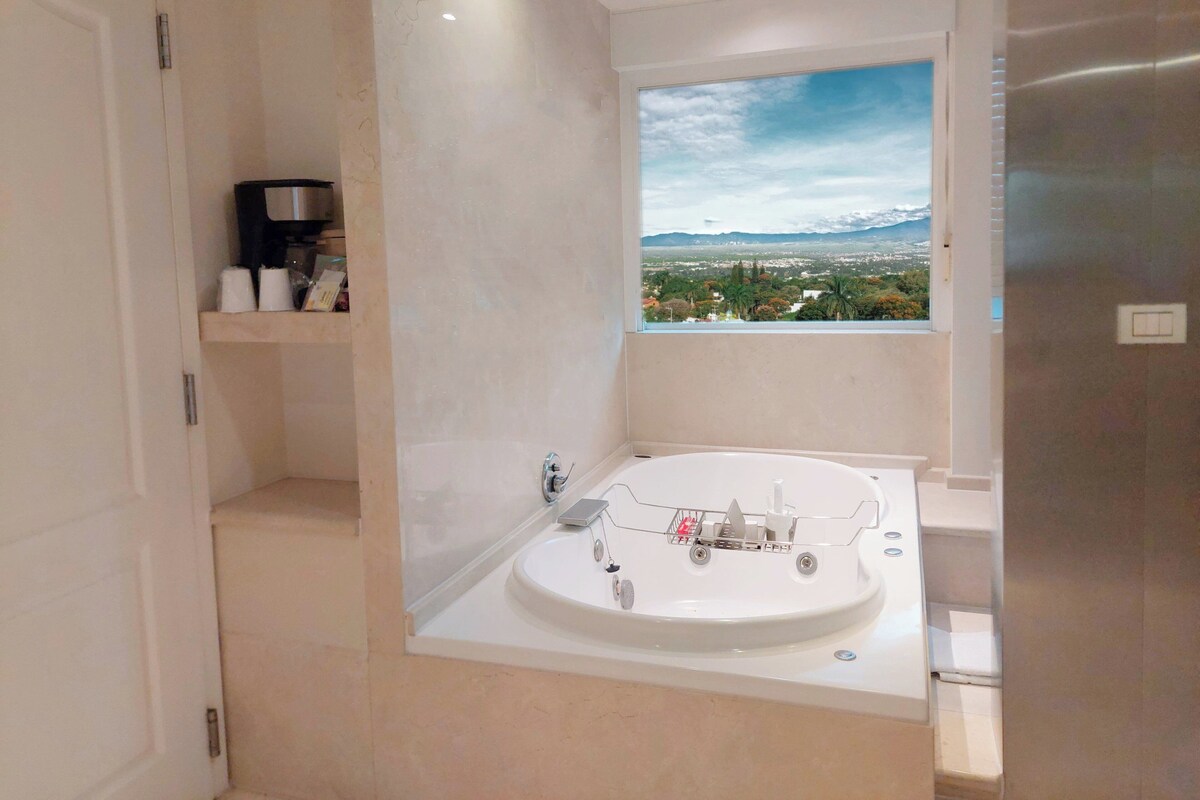 A modern bathroom with a large bathtub, a window with a cityscape view, and a shelf with toiletries.