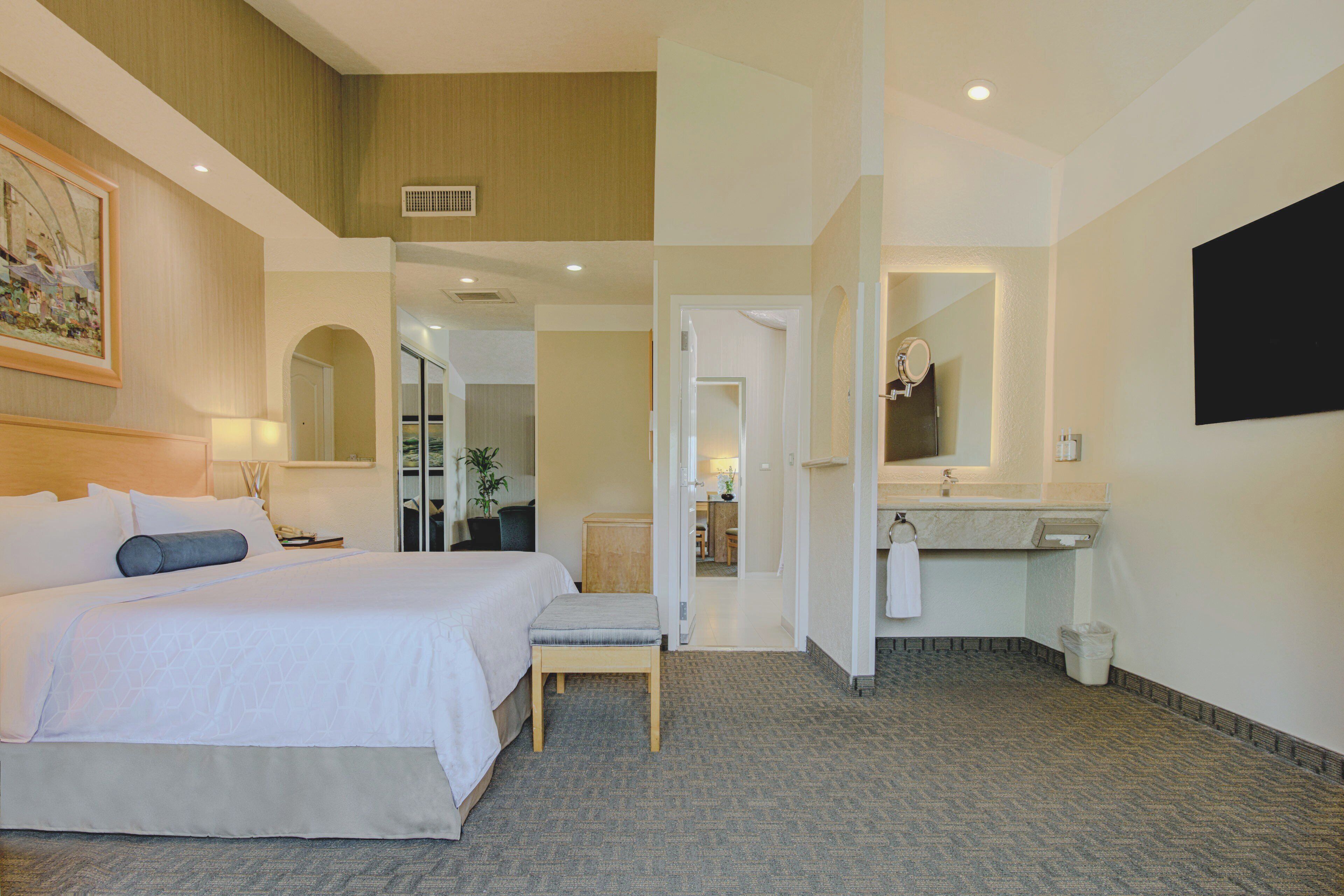 Habitación de hotel con una cama grande, un área de descanso, un baño con lavamanos y espejo, y un pasillo que da a otras habitaciones.