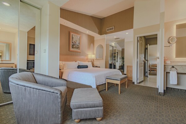 A hotel room with a bed, a sitting area with a chair and ottoman, a bathroom with a sink and mirror, and a hallway leading to other rooms. - Holiday Inn Cuernavaca by IHG (Cuernavaca)