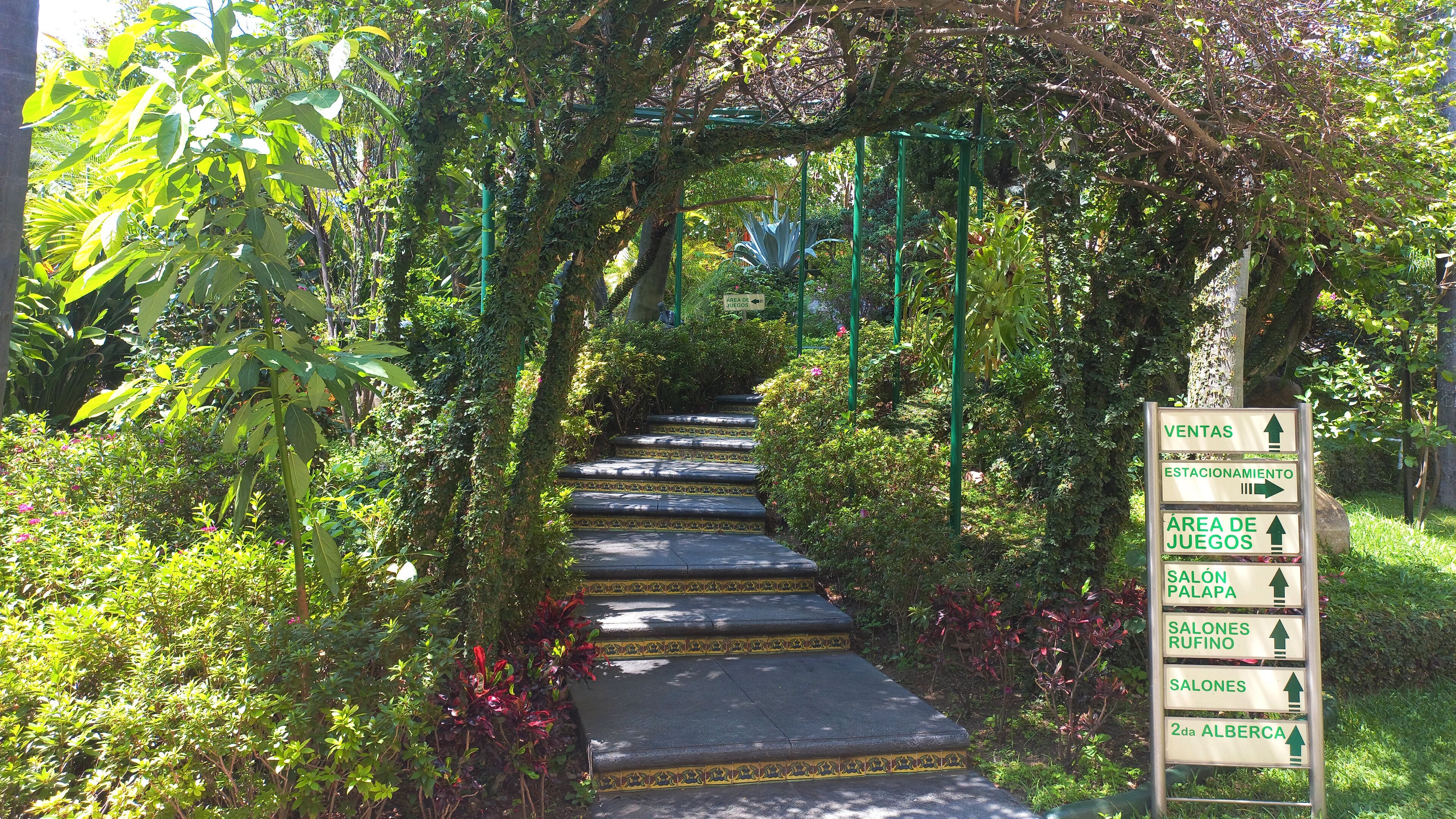 Un escalier entouré d’une végétation luxuriante, menant à un panneau indicateur indiquant la direction de différents lieux.