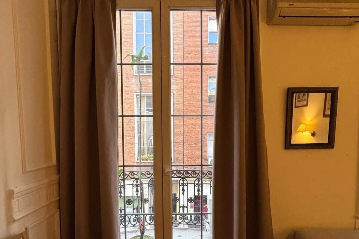 A room with a wooden herringbone floor, a bench, a bed, a window with curtains, and an air conditioning unit on the wall.
