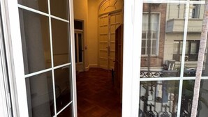 A hallway with a wooden floor, a glass door with a grid pattern, and a view of a building with multiple balconies. - Hotel Frossard (Buenos Aires)