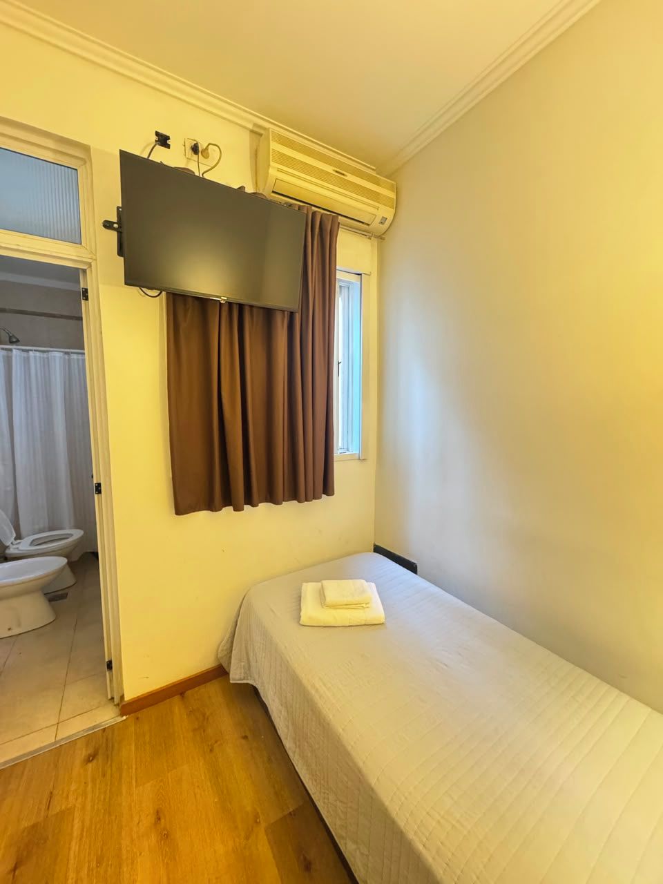 Una habitación pequeña con una cama, un televisor de pantalla plana montado en la pared, una ventana con cortinas y una puerta que da a un baño.