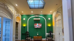 A room with a green wall, wooden flooring, a TV mounted on the wall, and two chairs. - Hotel Frossard (Buenos Aires)