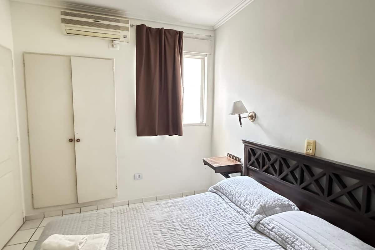 A neatly made bed with a quilted white duvet, a window with brown curtains, a bedside table with a lamp, and a wardrobe with a mirror.