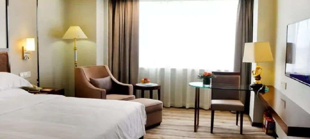 A hotel room with a bed, two armchairs, a small table, a TV, and a window with curtains.