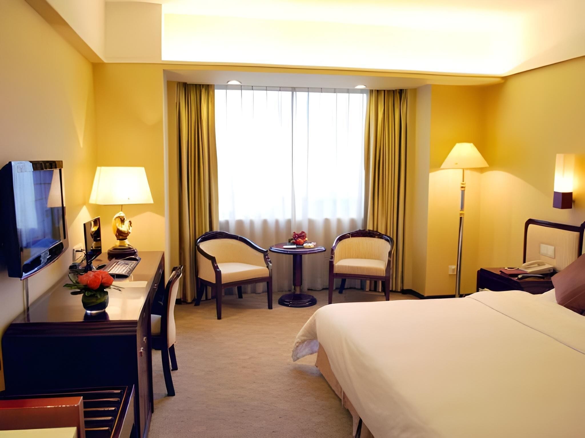 A hotel room with a bed, a desk, two chairs, a television, a lamp, and a window with curtains.