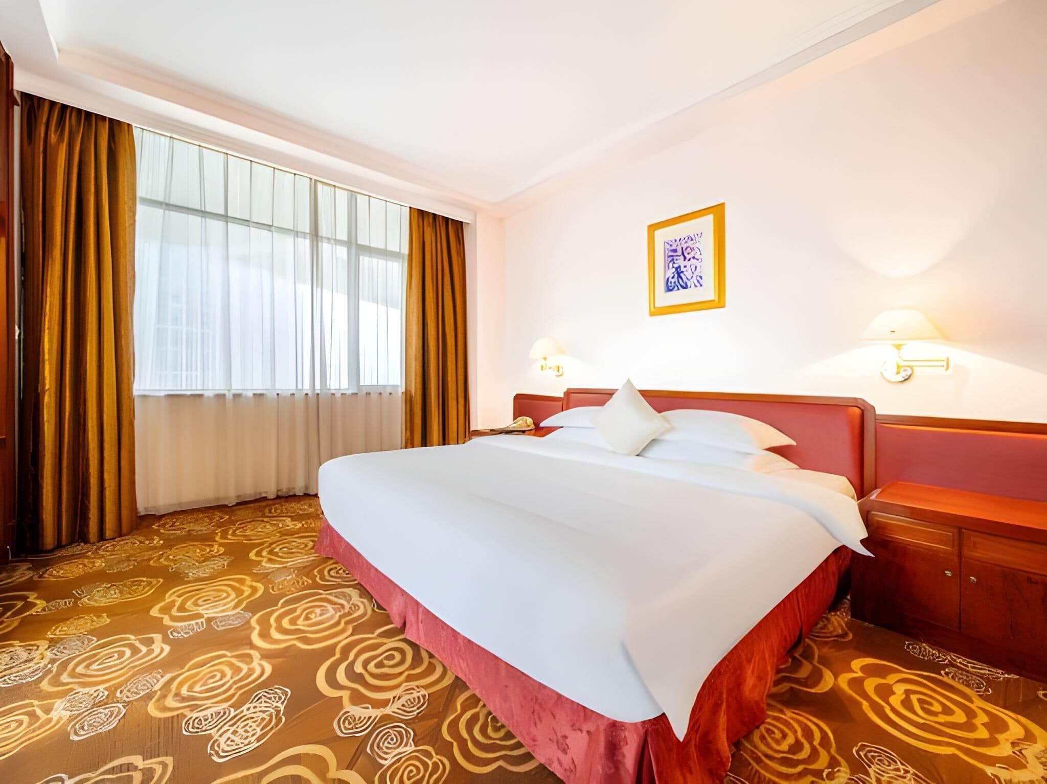 A hotel room with a large bed, a window with curtains, a framed picture on the wall, and a patterned carpet.