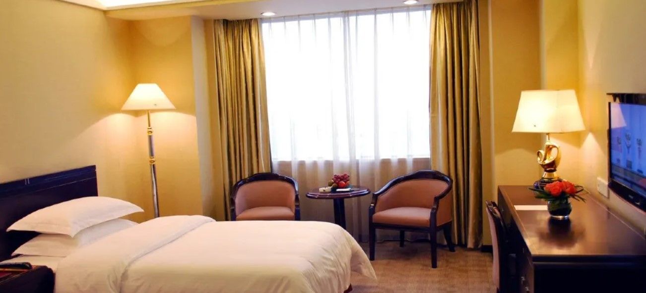 A hotel room with a bed, two armchairs, a small table with a vase, and a TV on a wooden table.