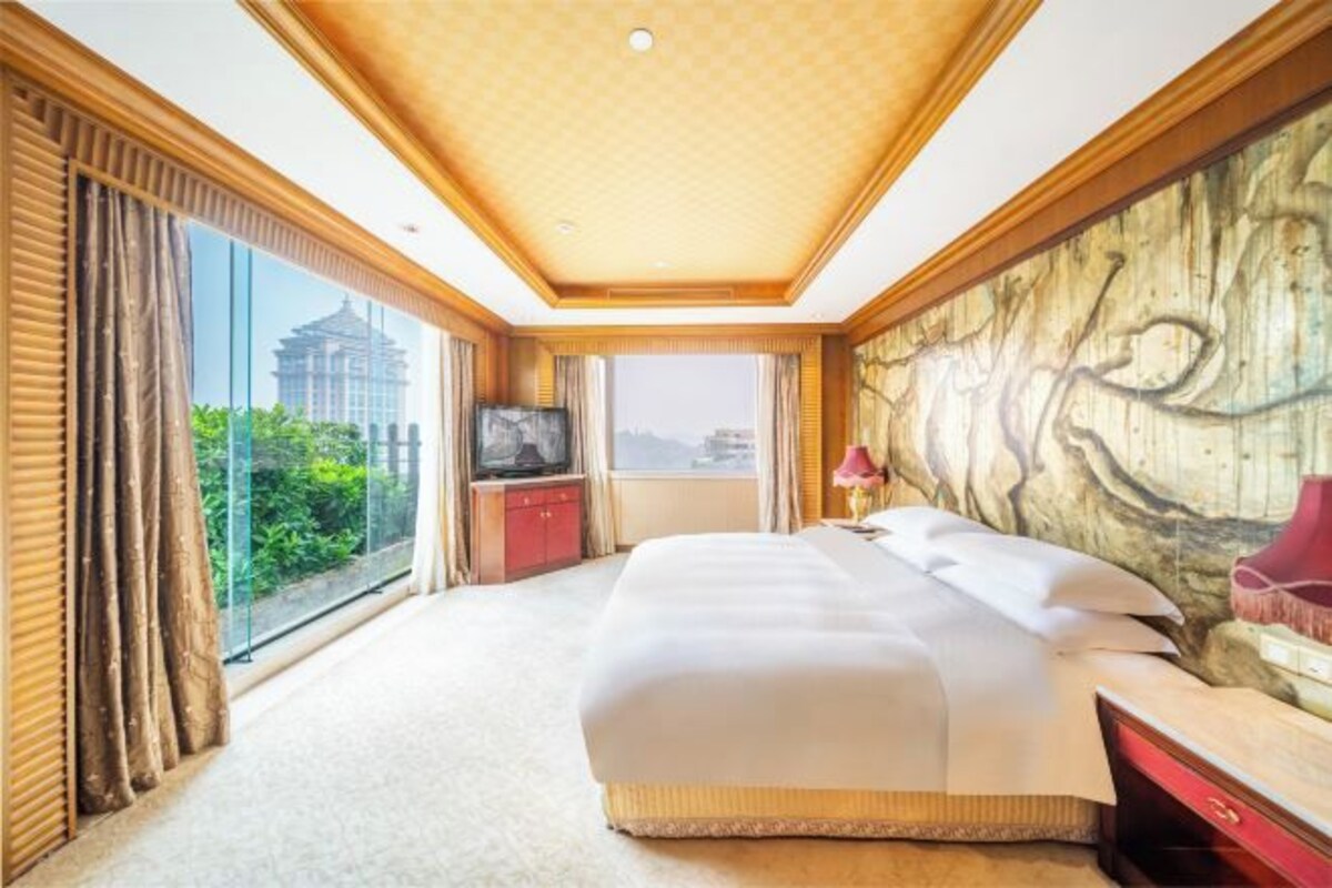 Habitación de hotel con una cama grande, televisión, escritorio y vista a un edificio a través de amplias ventanas.