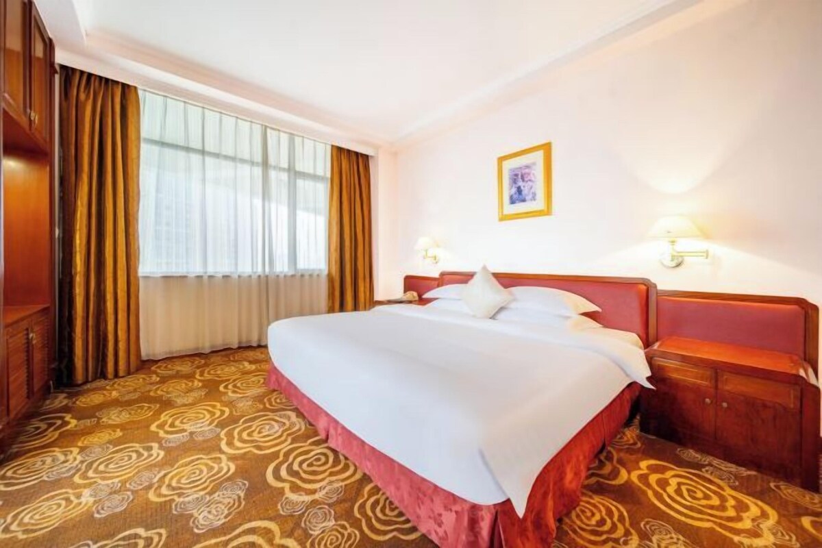 Habitación de hotel con una cama grande, cabecera de madera, dos lámparas de noche, un cuadro enmarcado en la pared y una alfombra estampada.