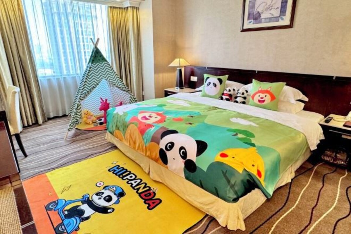 Una habitación de hotel con una cama que tiene una colcha colorida con estampado de panda, una pequeña tienda verde y blanca a rayas, y una alfombra amarilla con una ilustración de panda.