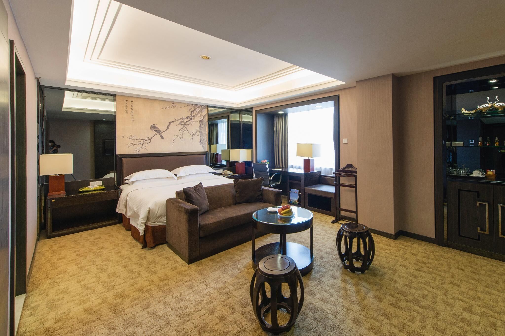 Quarto de hotel com uma cama grande, um sofá, uma mesa redonda pequena, dois banquinhos, uma escrivaninha e uma janela ampla.