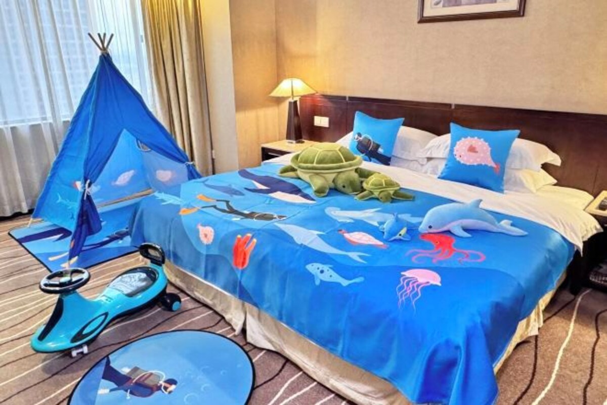 Habitación de hotel con una cama cubierta con una colcha azul con motivos marinos, un cojín con forma de tortuga verde y una estructura azul parecida a una tienda de campaña en el suelo.