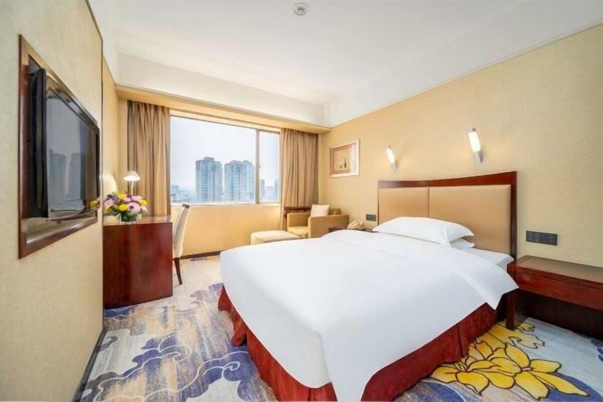 Habitación de hotel con una cama grande, un escritorio con lámpara, una silla, un televisor y vistas a edificios altos.