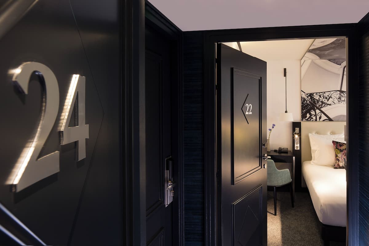 A modern hotel room with a dark theme, featuring a large number '24' on the wall, a bed with white linens, a desk with a lamp, and a small seating area.