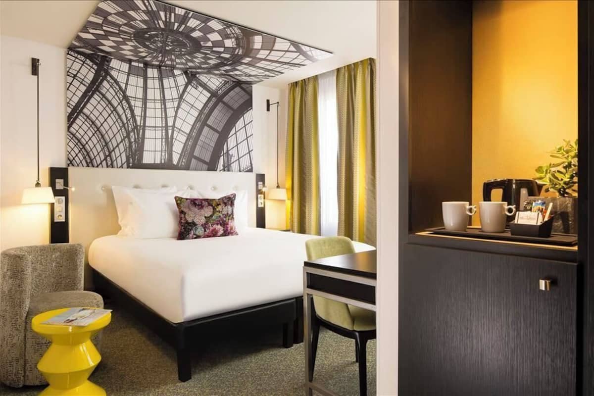 A hotel room with a bed, a desk, a chair, a small yellow stool, and a cabinet with cups and a plant.