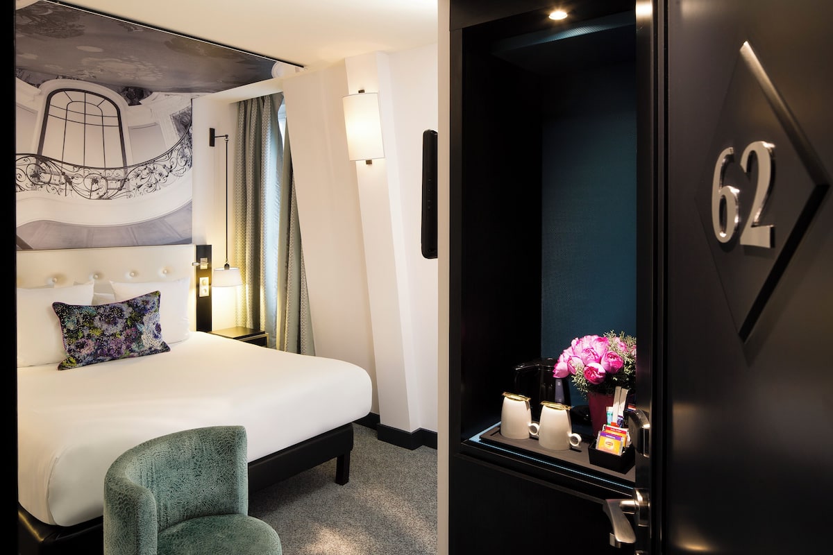 A hotel room with a bed, a green armchair, a nightstand with a lamp, a wall-mounted light, and a door with the number 62.