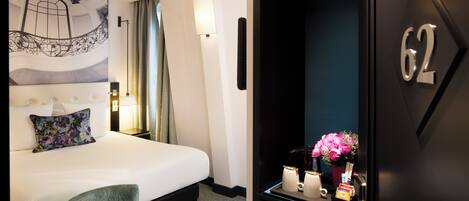 Habitación de hotel con una cama, un sillón verde, una mesita de noche con lámpara, una luz en la pared y una puerta con el número 62.