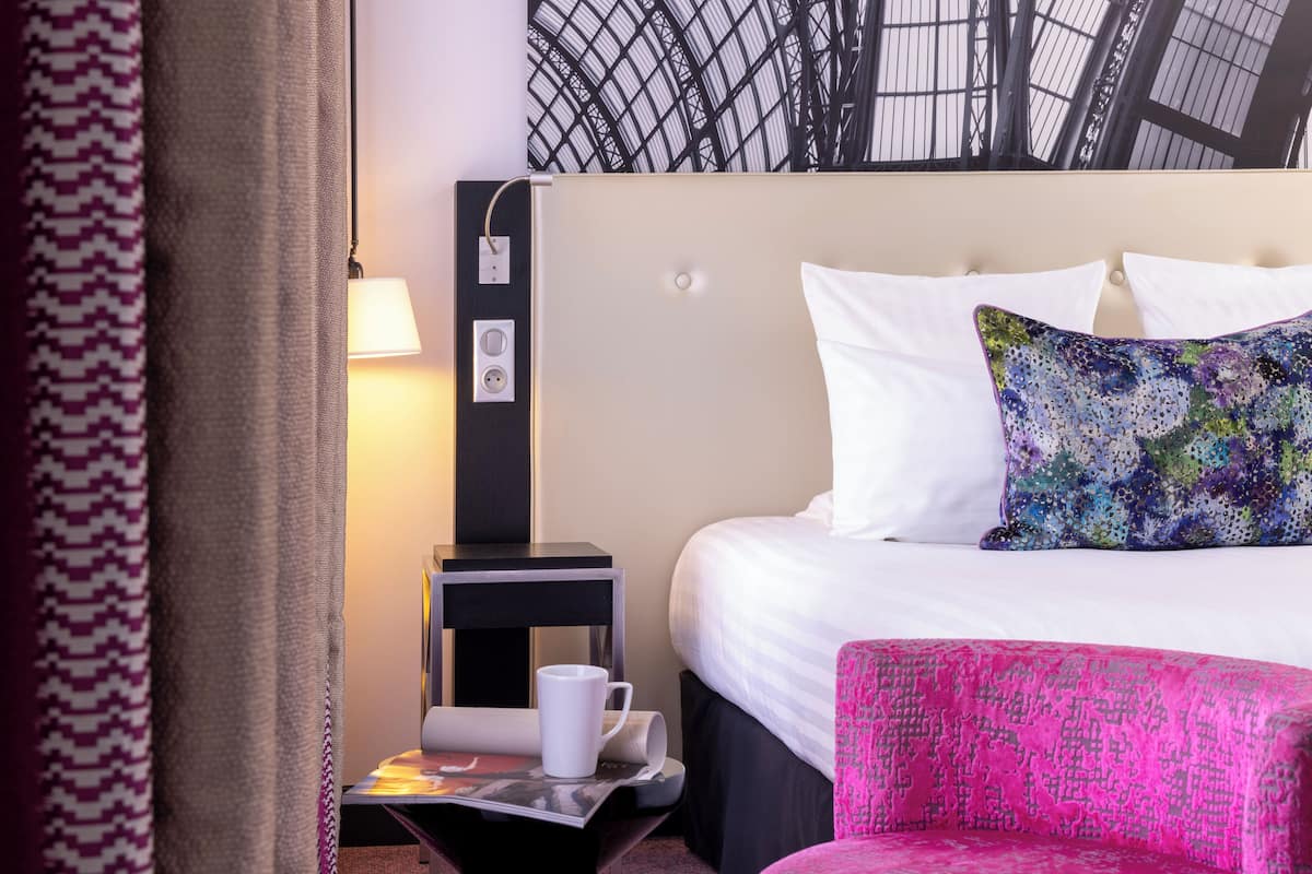 A hotel room with a bed, a nightstand with a lamp, a chair, and a patterned wall.