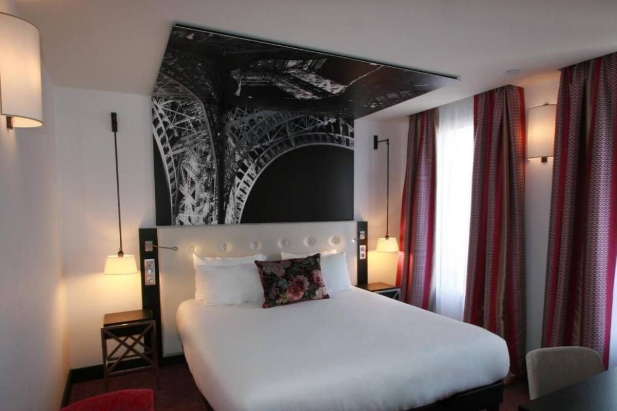 A hotel room with a large bed, a red chair, a desk, and a wall-mounted artwork of a bridge.