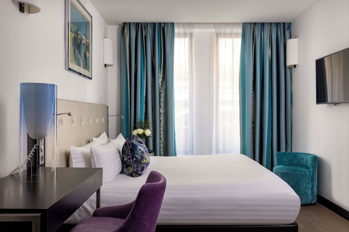 A hotel room with a bed, a desk with a lamp, a chair, and a TV on the wall.