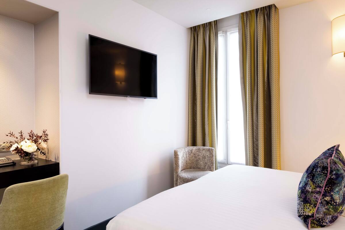 A hotel room with a bed, a TV mounted on the wall, a desk with a lamp, and a chair.