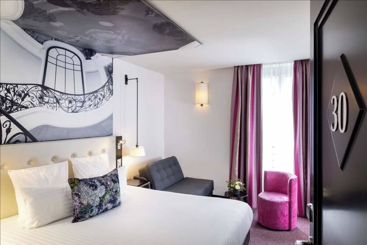 A hotel room with a bed, a desk, a chair, a wall-mounted artwork, a lamp, and curtains.