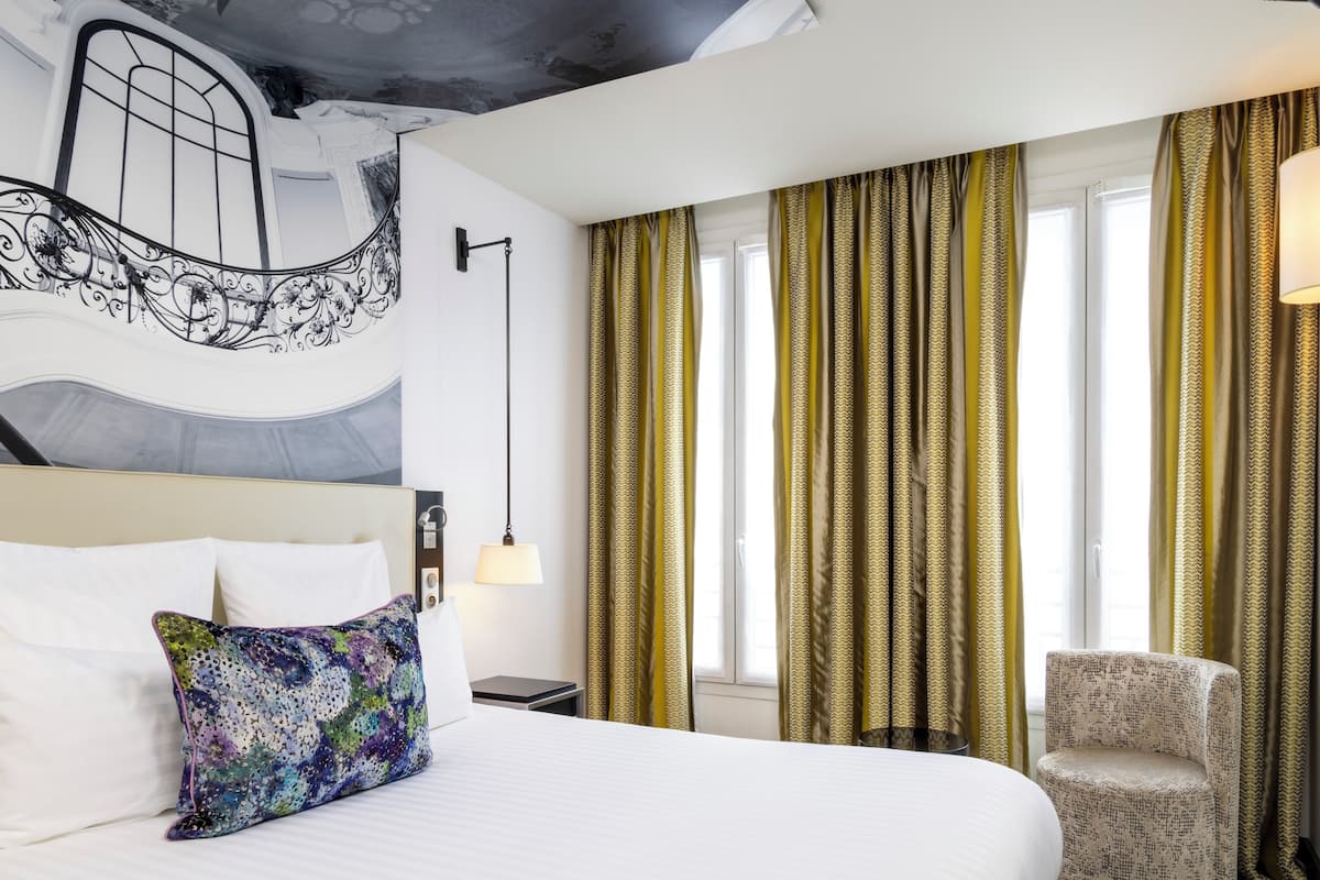 A hotel room with a bed, a chair, a lamp, a window with curtains, and a wall with a decorative design.